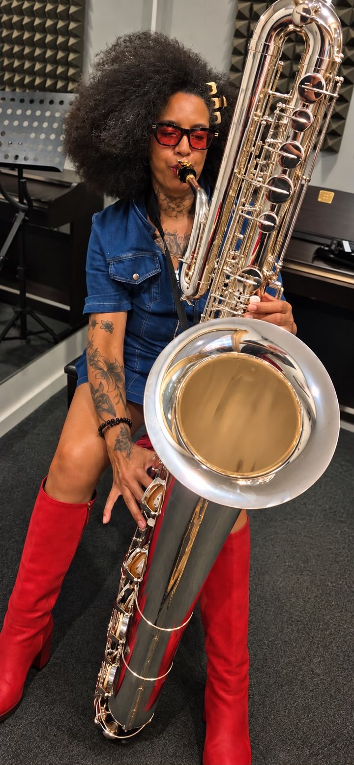 Rosemary Quaye seated with her full-length silver bass saxophone, wearing red knee-high boots